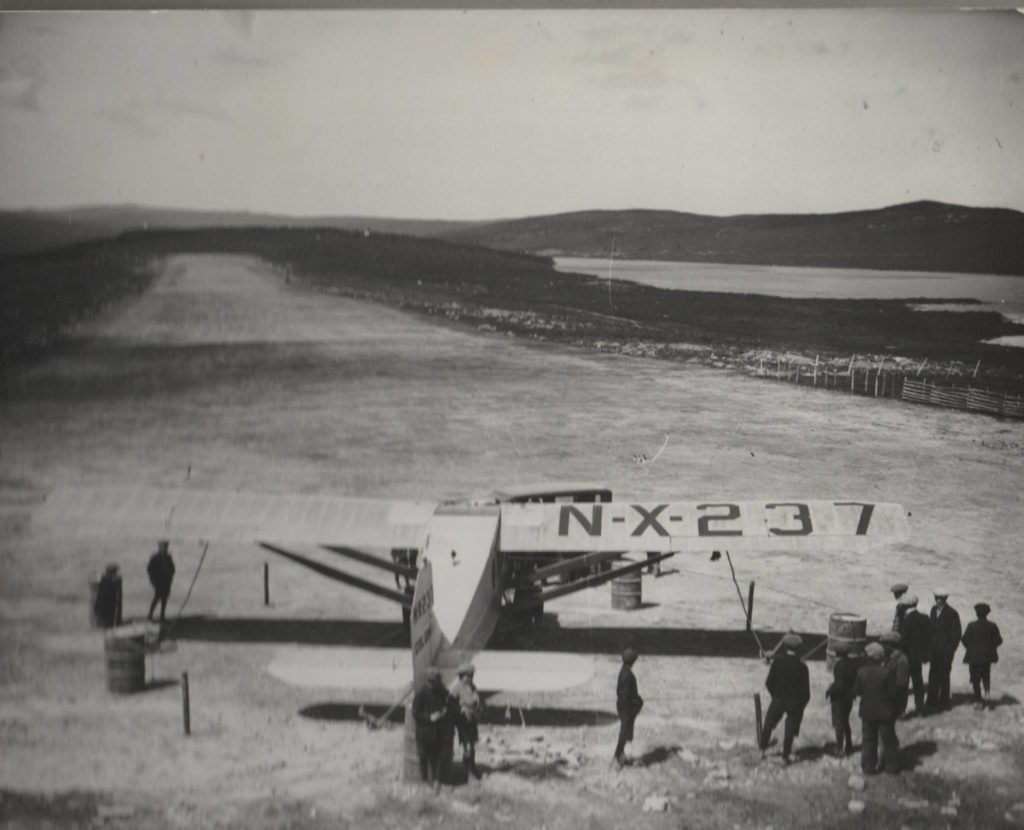 The aircraft Columbia looking down the stretch of the Harbour Grace runway with Lady Lake in the background. A group of people are milling around the aircraft which is tied down.