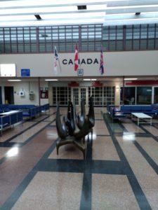 The inside of the Gander International Airport Terminal. Visible is the statue "Birds of Welcome". On the back wall is the word Canada and the Newfoundland flag, the Canadian flag, and the Union Jack.