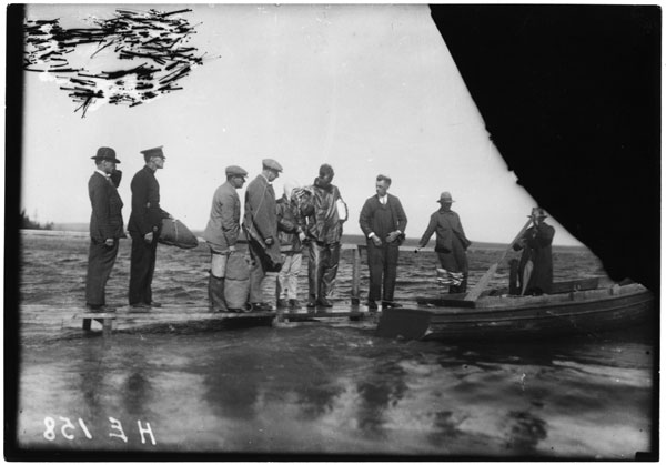 Charles and Anne Morrow Lindberg on a wharf. Six men are on the wharf with them, and a seventh man is in a small boat. The view is mostly of water, but part of the image is scratched in the top left corner and black in the top right corner.