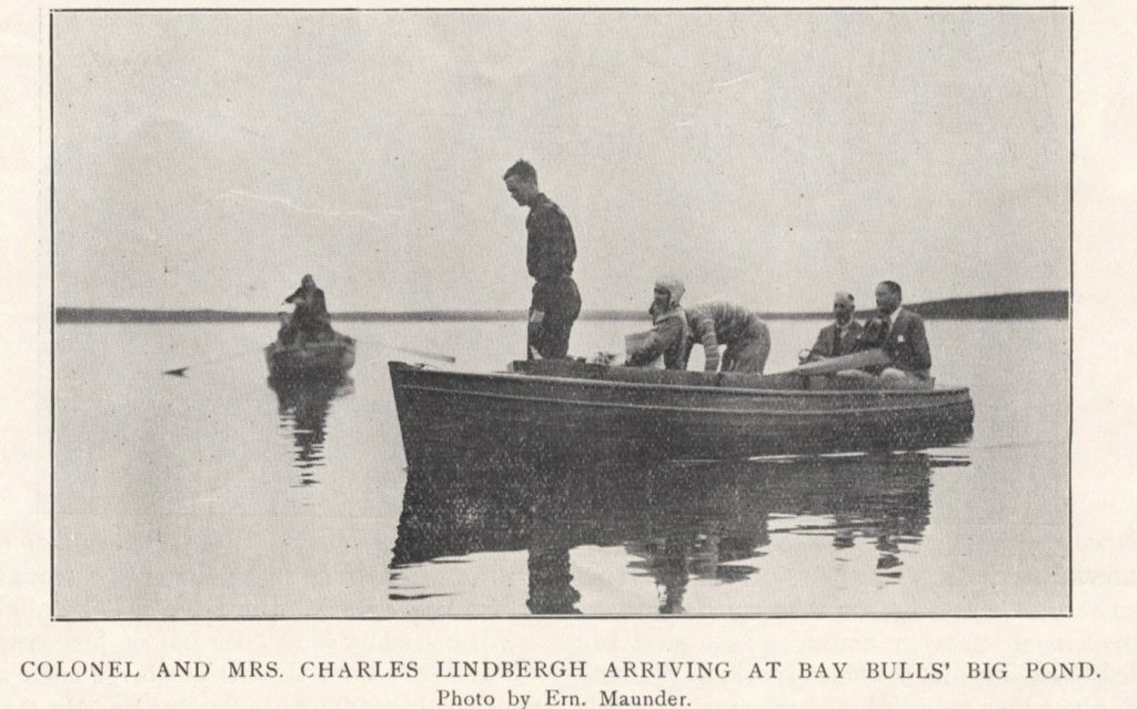A black and white photo of two small boats on a calm pond. The background boat has someone rowing. The foreground boat has a man standing in the front, two men in suit jackets sitting in the rear, and Ann Lindbergh sitting in the middle, with Charles next to her, looking as if he is about to sit. The caption reads Colonel and Mrs Charles Lindbergh arriving at Bay Bulls Big Pond. Photo by Ern Maunder