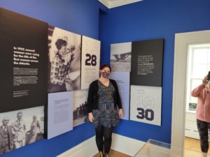 A woman in a blue dress and black sweater stands in a corner next to two groups of exhibit panels. Next to her, very clearly, is a picture of Amelia Earhart.