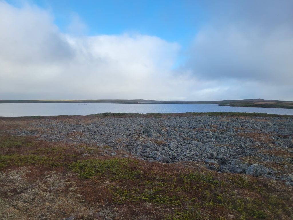 A view of barrens and a pond under a blue, cloudy sky.